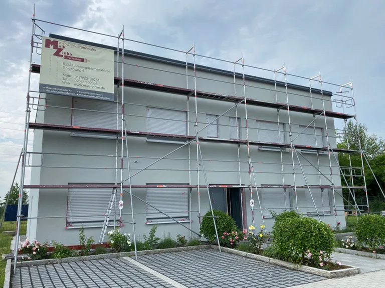 Mehrfamilienhaus im Gerüst mit grau gestrichener Fassade, davor ein gepflegtes Rosenbeet